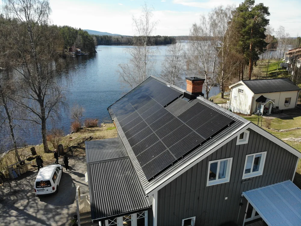Grått hus med ett stort tak av helsvarta solpaneler vid en sjö i Järvsö, installerat av Håpe Kraft